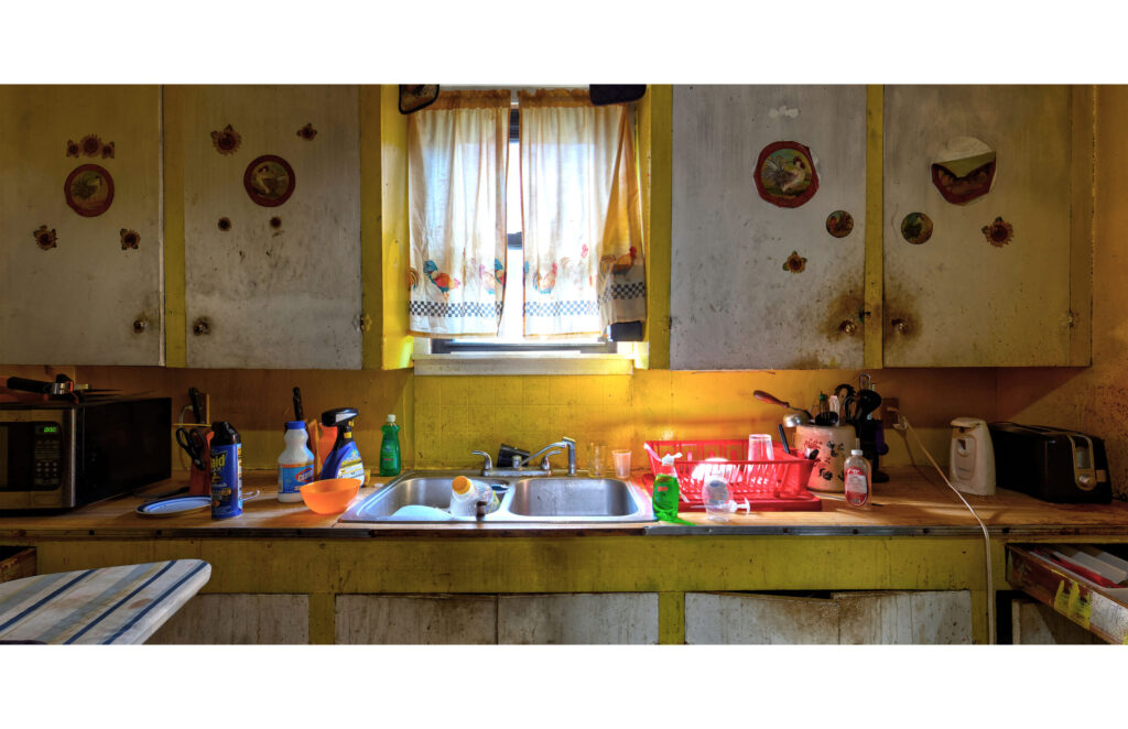 Yellow kitchen with rooster curtains, worn cabinets, sink, dishes, cleaning supplies, microwave, and ironing board.