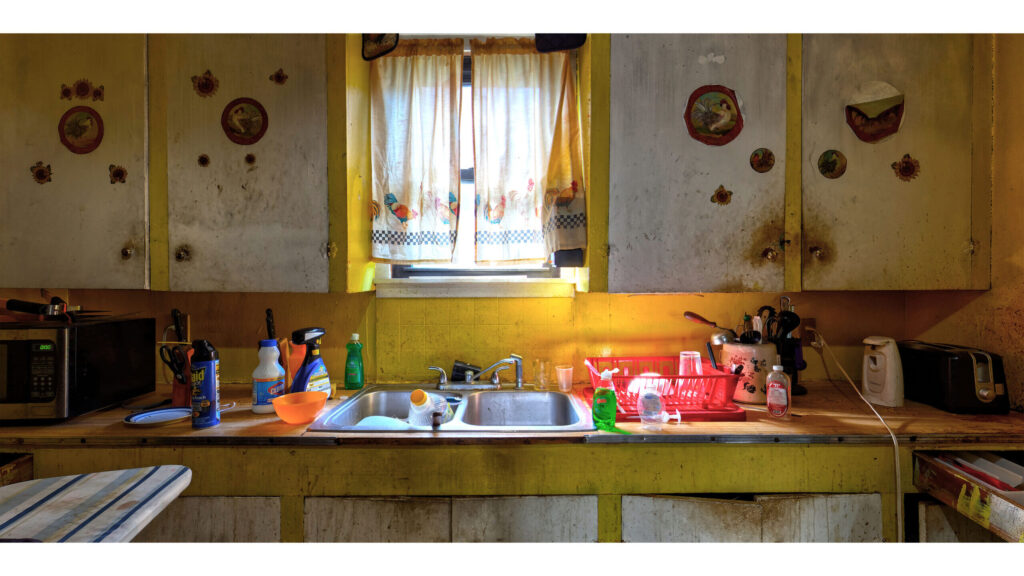 Kitchen with worn cabinets, curtained window, sink, and household items across the counter.