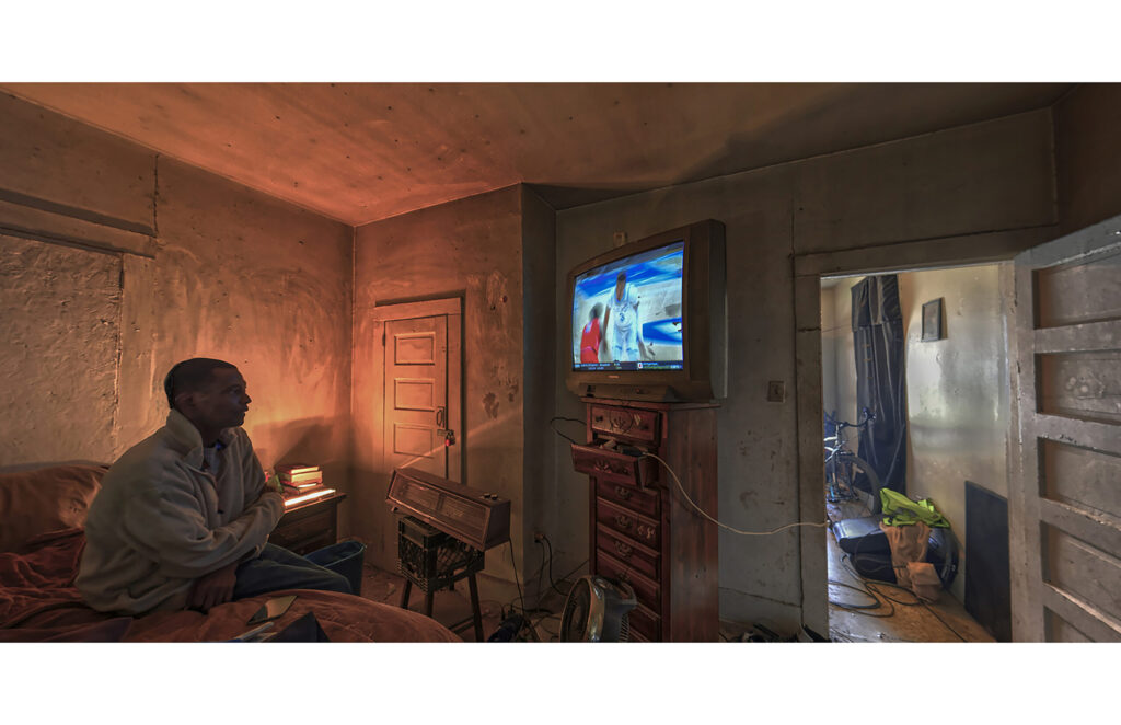 Man seated in a dim room watching television beside an open doorway.