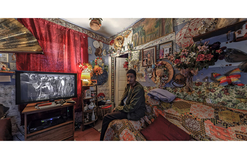 Woman seated in a richly decorated living room filled with artwork, textiles, flowers, and personal objects.