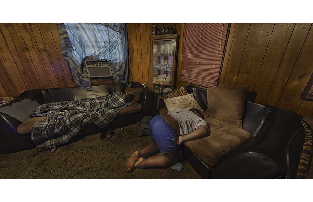 luttered wood-paneled room with bed, piled clothing, furniture, and household belongings.