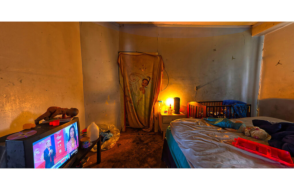 Dim bedroom with bed, crib, television, glowing lamp, and fabric covering a doorway.