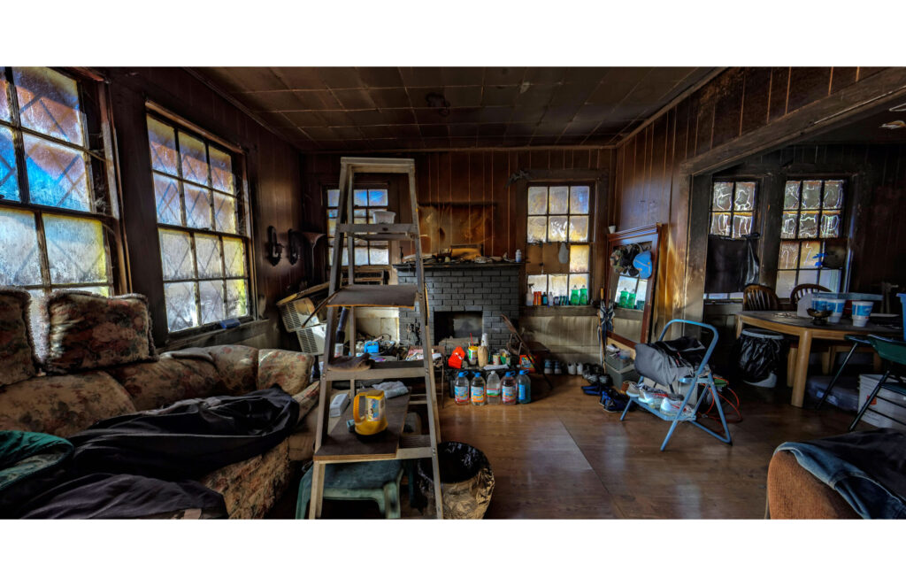 Large dim wood-paneled room crowded with furniture, ladder, windows, and household objects.