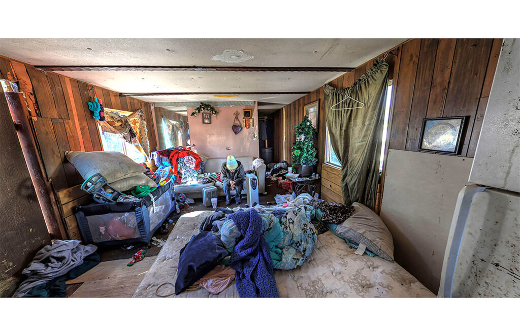 luttered wood-paneled room with bed, piled clothing, furniture, and household belongings.