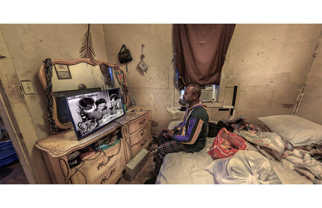 Person seated on a bed watching television in a worn bedroom filled with personal belongings.
