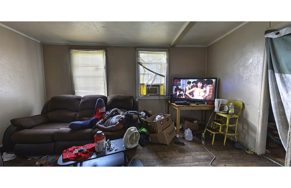 Person reclining on a sofa in a sparsely furnished living room with television and two windows.