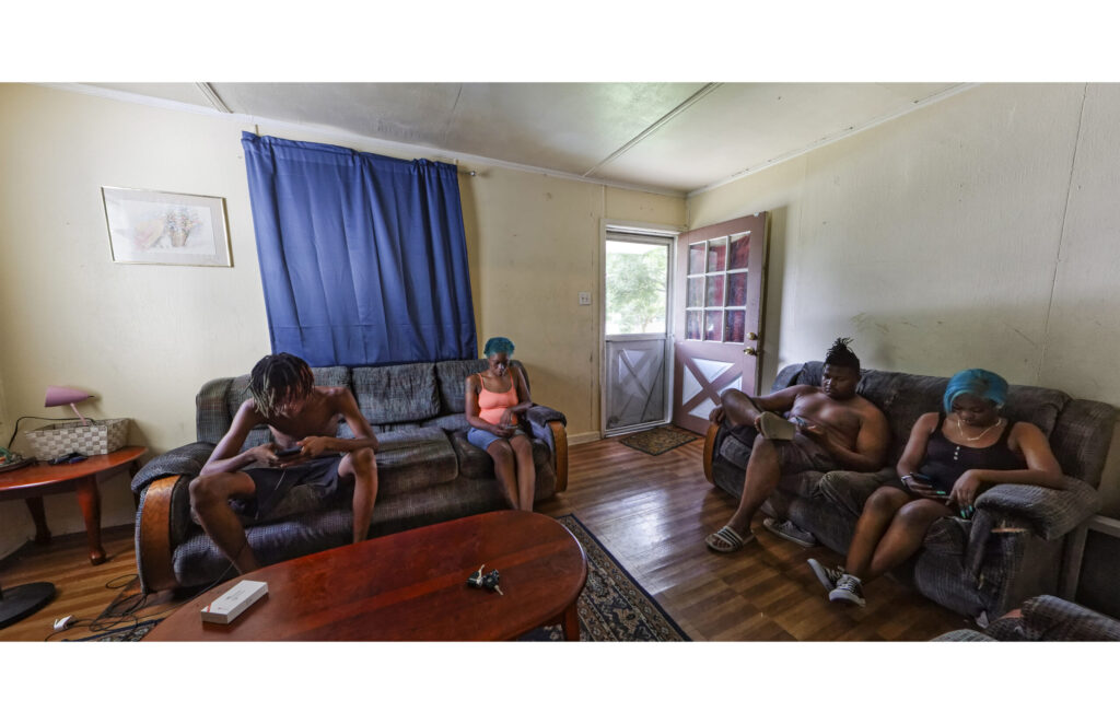 Four young people seated on sofas in a living room with blue curtains and open front door.