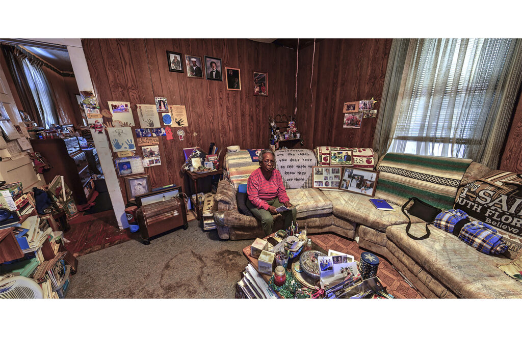 Older woman seated in a wood-paneled living room surrounded by photographs, books, and personal keepsakes.
