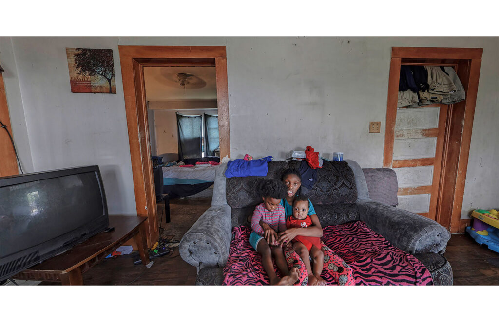 A young woman is seated with two young children in a living room with a television and an adjoining bedroom.