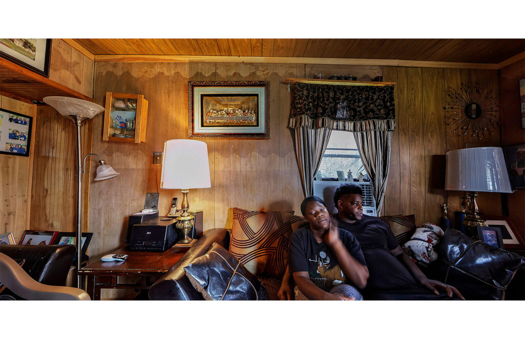 Two people seated in a wood-paneled living room with lamps, framed pictures, and curtained window.