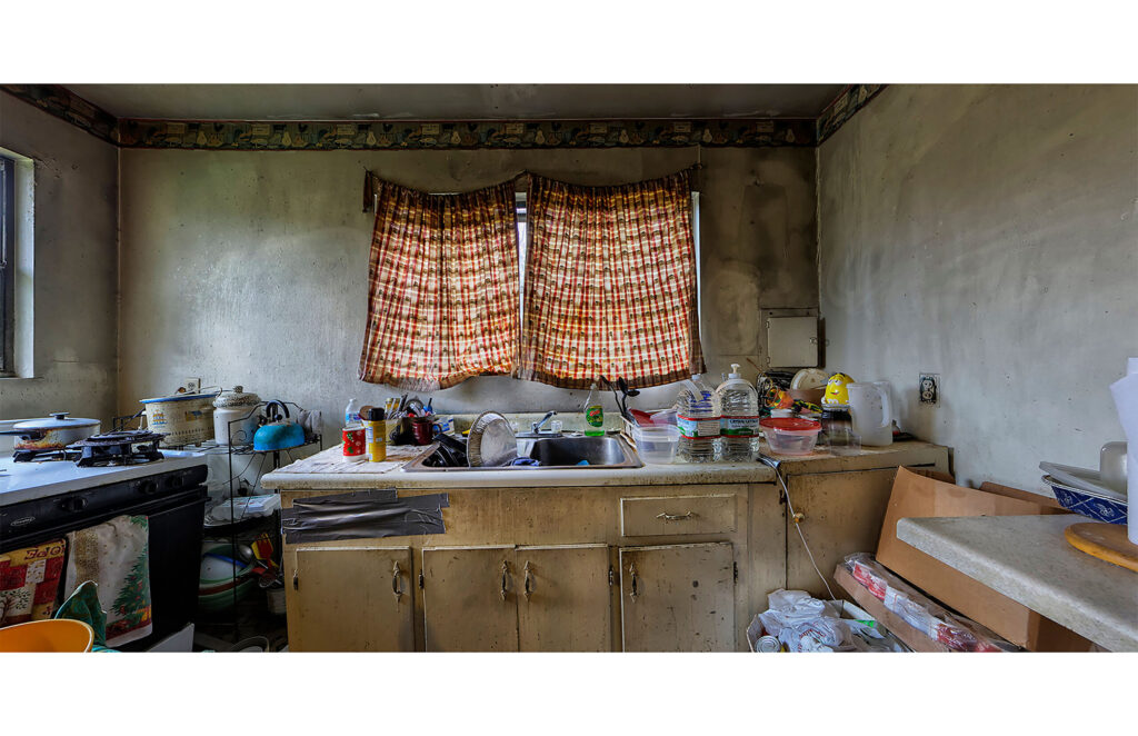 Worn kitchen with plaid curtains, sink filled with dishes, pots on the stove, water bottles, containers, and aging cabinets.