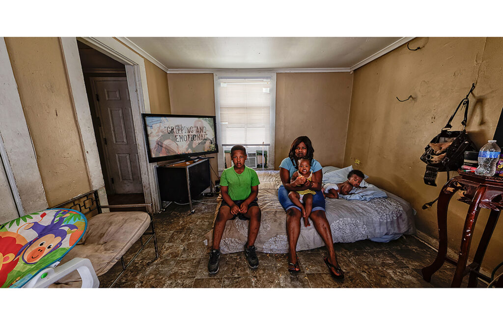 Woman seated on a bed with three children in a sparsely furnished room.